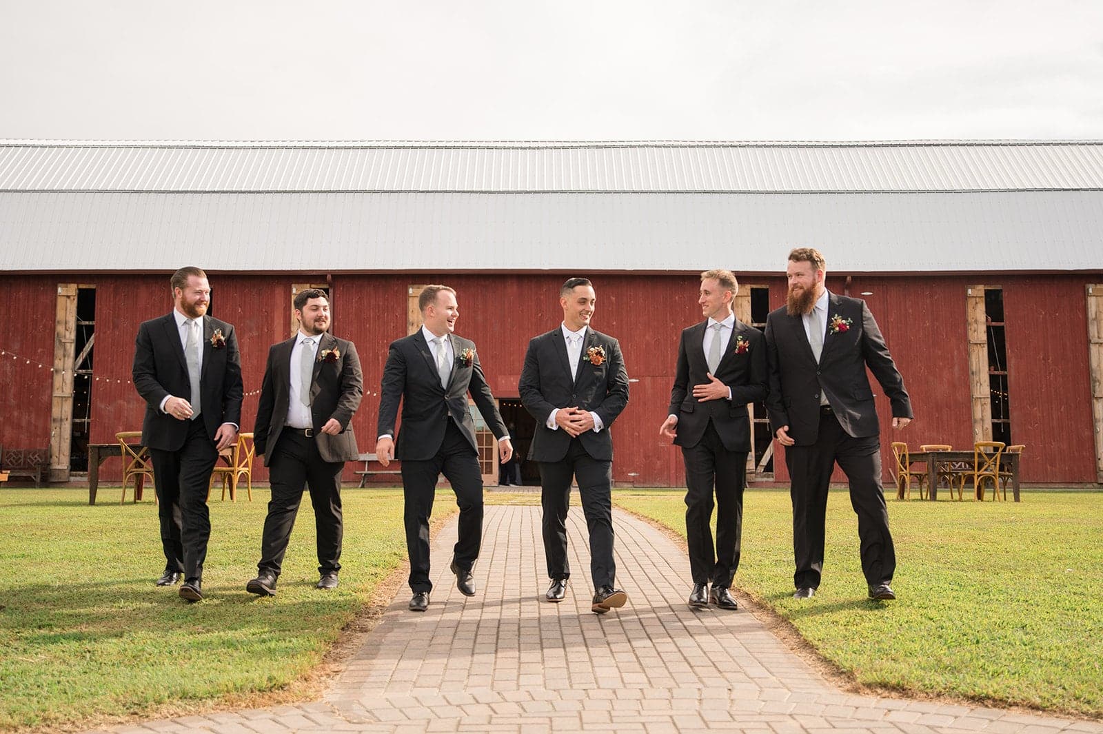A group of six men in suits walks confidently along a path in front of a rustic barn.