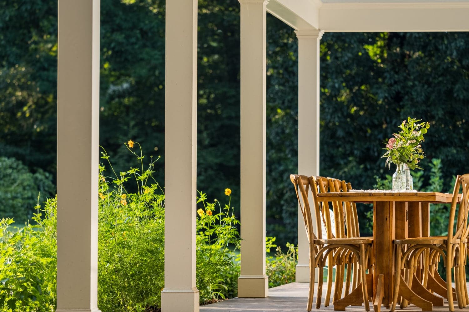 A wooden dining table is set on a porch surrounded by lush greenery.