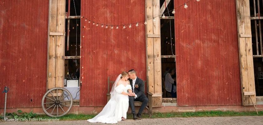 A bride and groom sit together outside the red barn at Honeys Harvest Farm with string lights and a wooden wagon wheel nearby.