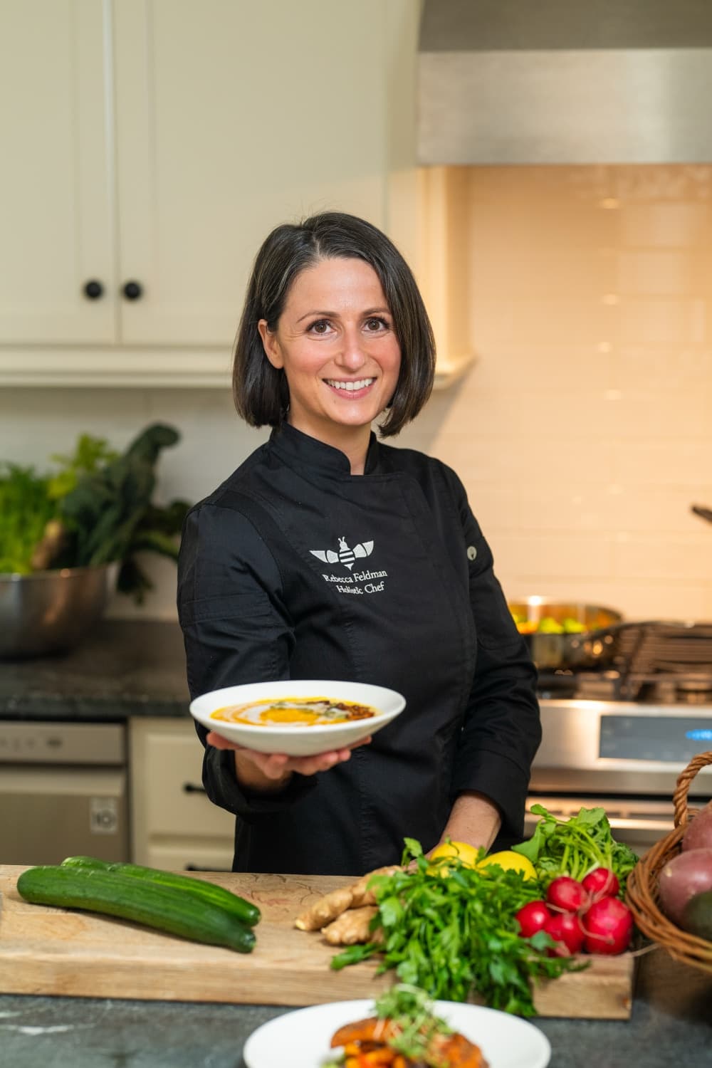 A smiling chef in a black coat holds a plate of food in a kitchen filled with fresh vegetables.