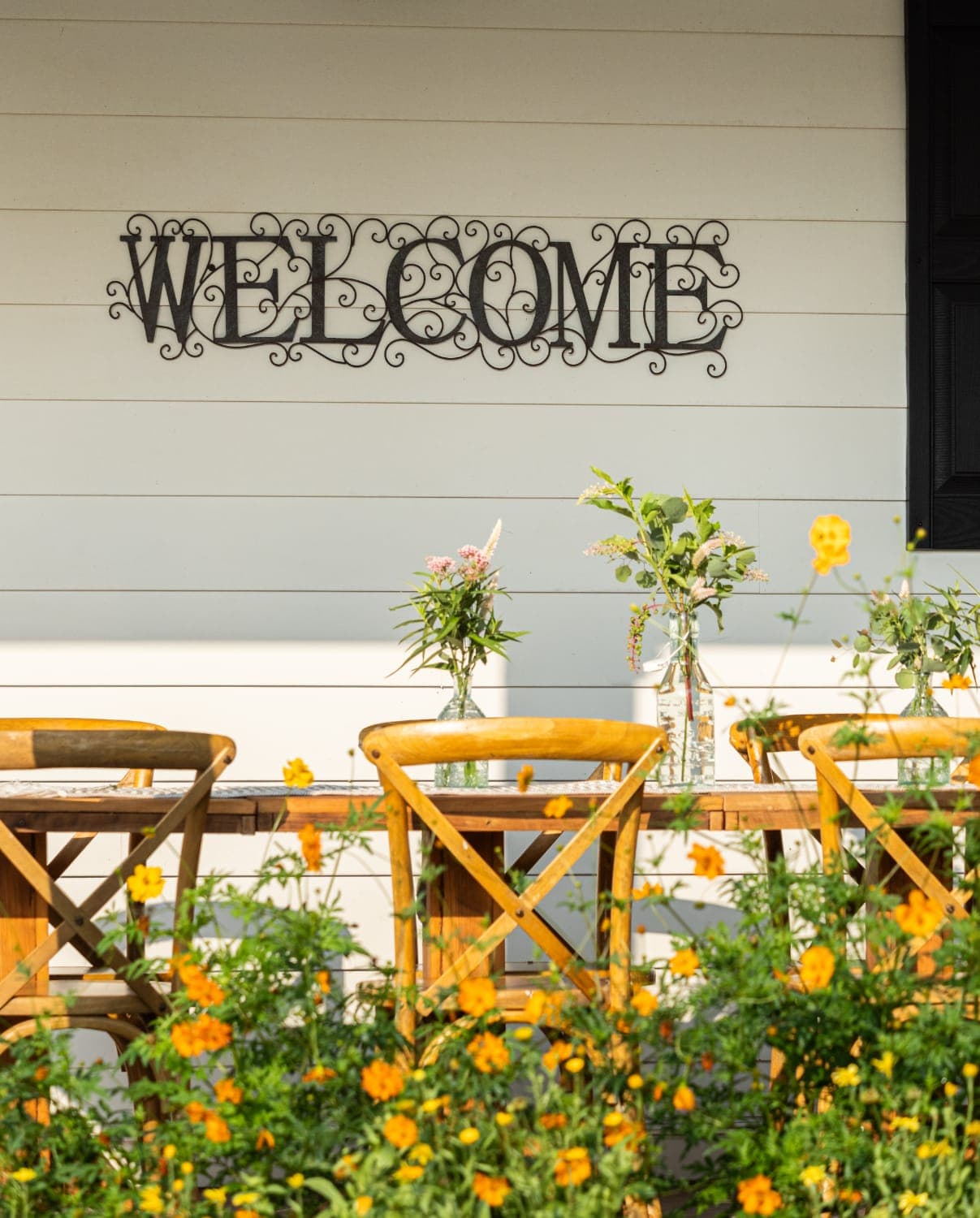 A "WELCOME" sign hangs above a rustic table with floral arrangements, surrounded by vibrant yellow flowers.