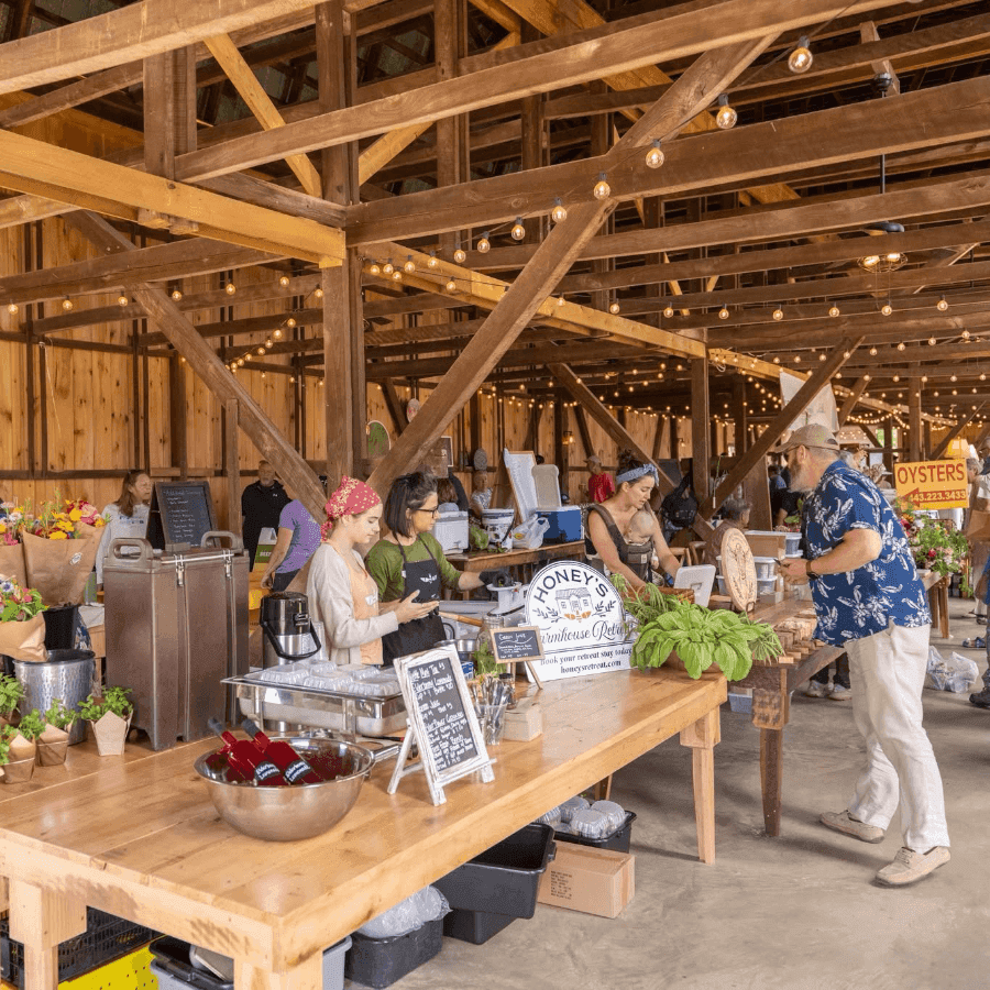 A lively farmers' market scene inside a rustic barn, with vendors and customers interacting among wooden beams and string lights.