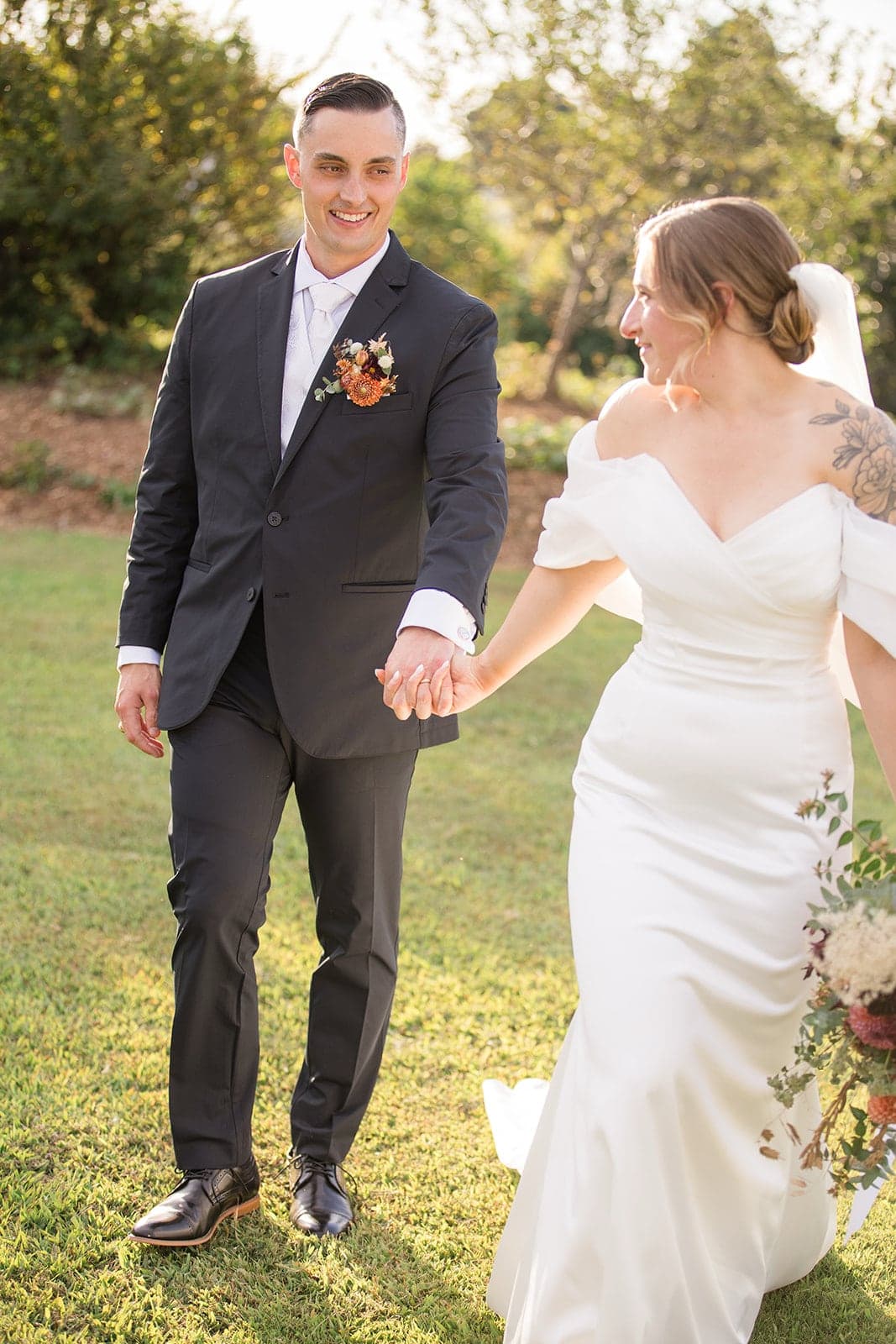 A bride and groom hold hands while walking in a sunlit outdoor setting.