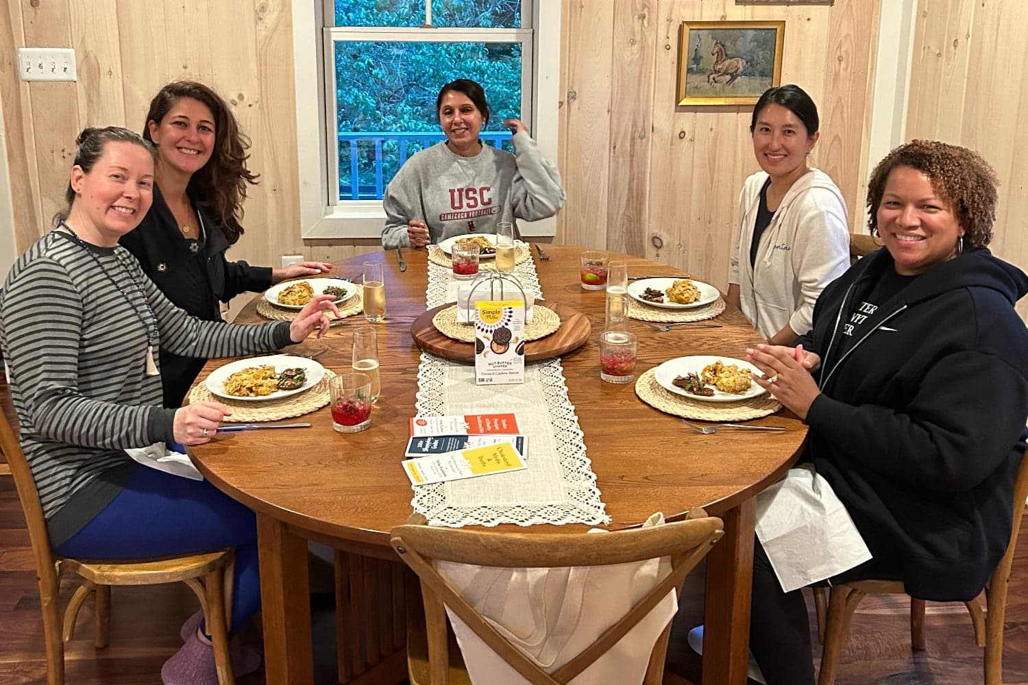 A group of five women sit around a dining table, enjoying a meal and drinks in a cozy setting.