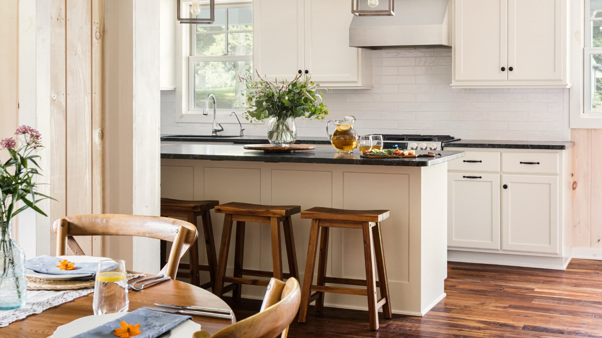 A bright, modern kitchen with white cabinetry, a dark countertop, and a dining area featuring wooden chairs and a vase of flowers.