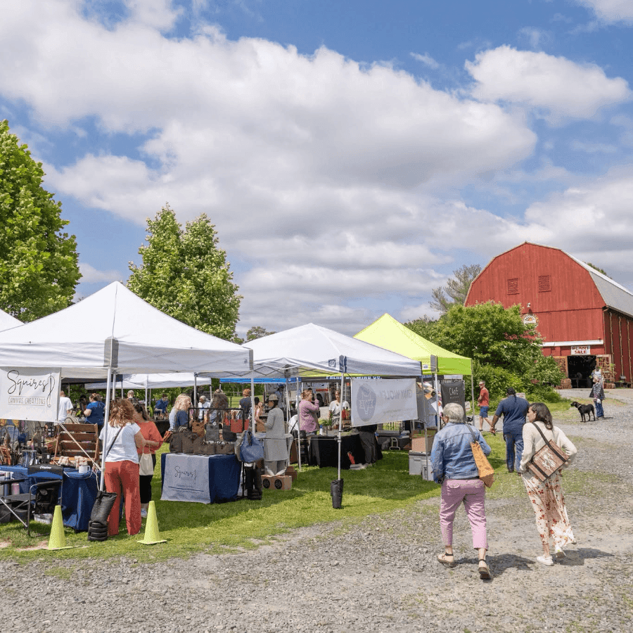 A bustling outdoor market with vendor tents, visitors, and a red barn under a partly cloudy sky.