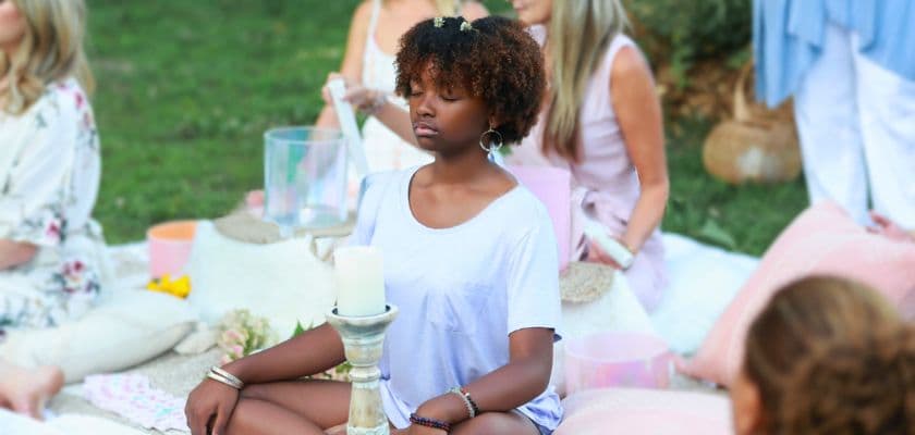 A woman with curly hair sits in meditation amid a group on a grassy area, surrounded by soft decor and candles.