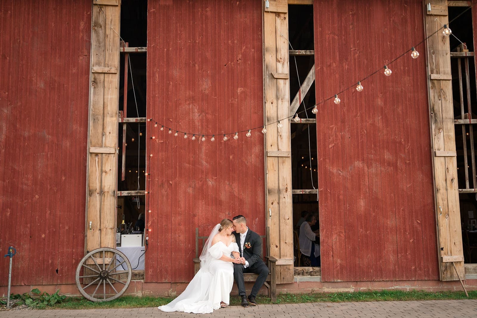 A couple sits together, sharing a tender moment in front of a rustic red barn decorated with string lights.