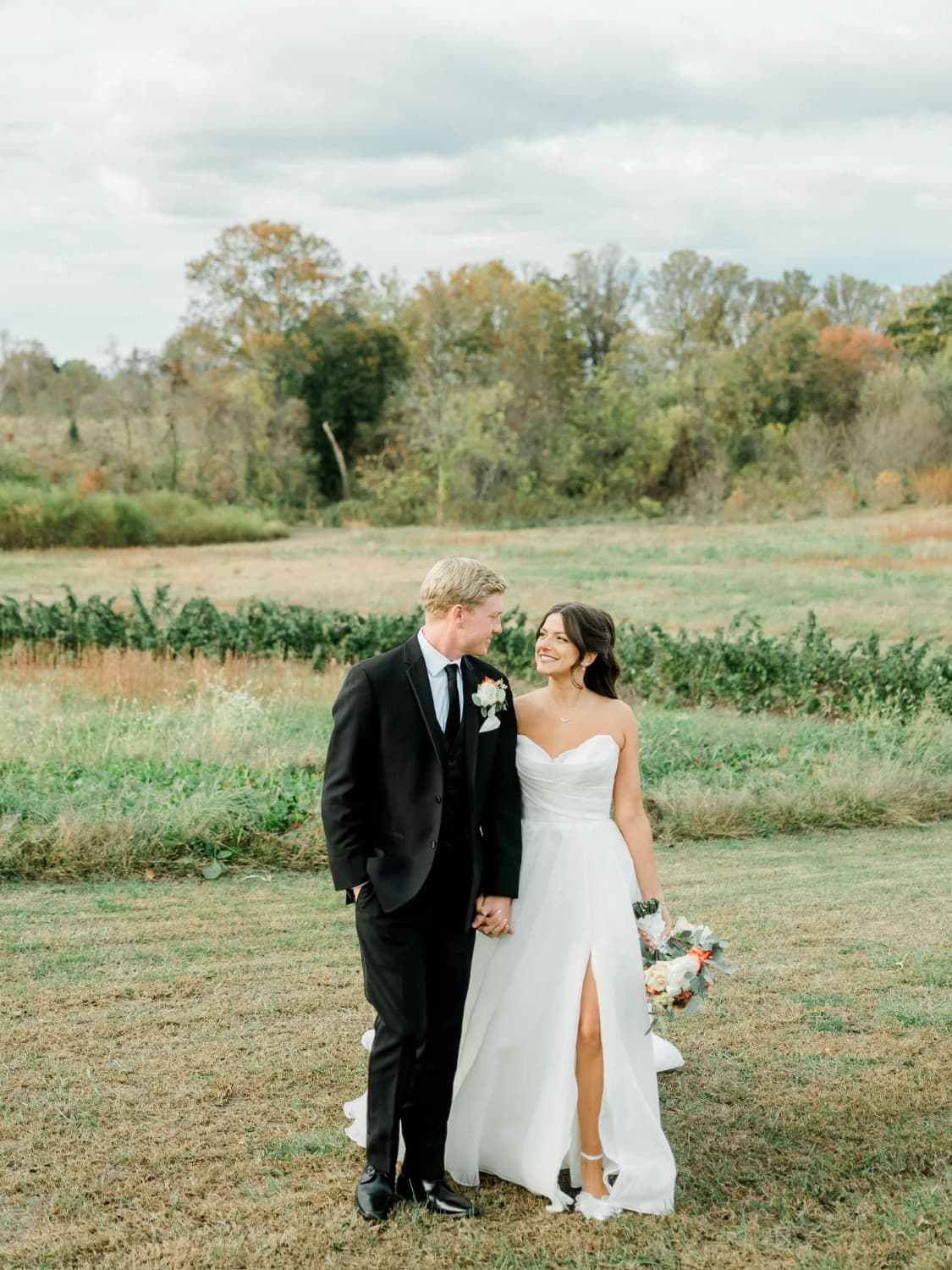 A smiling couple holds hands while walking in a scenic outdoor setting, surrounded by greenery and fields.