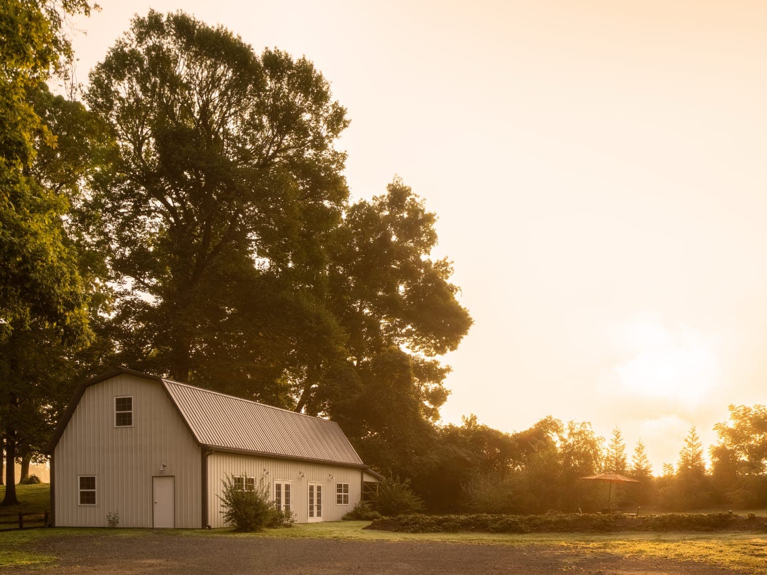 A white barn with a metal roof is surrounded by trees at sunset.