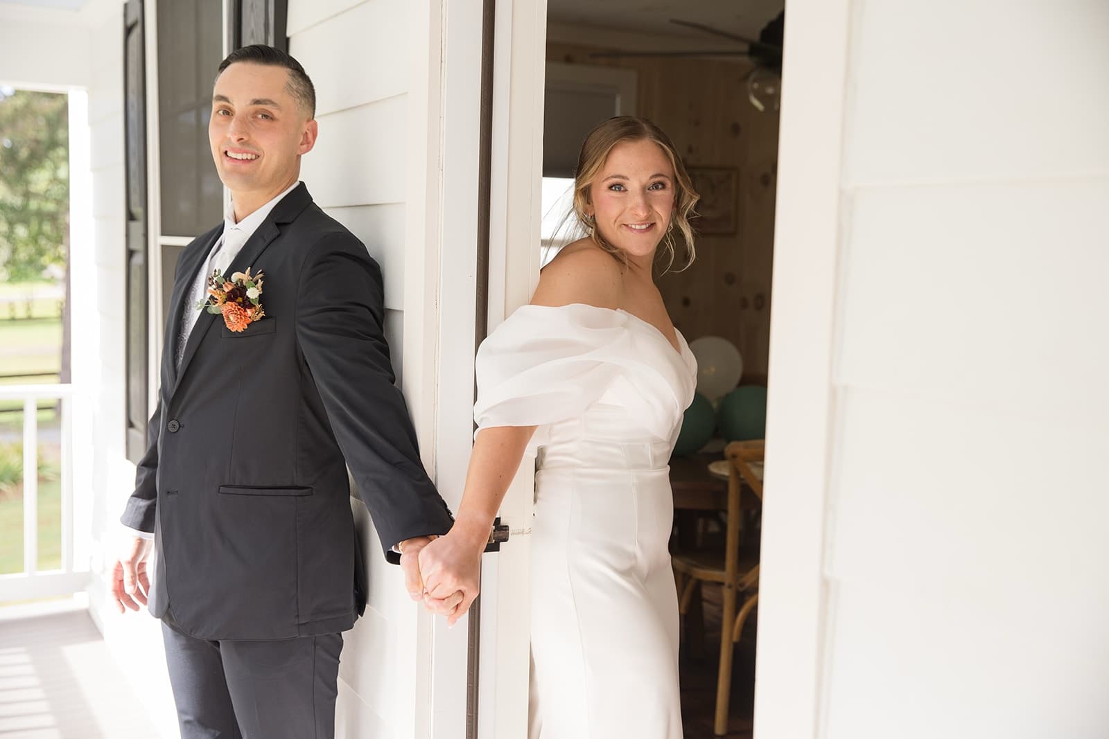 A couple in formal attire holds hands at a doorway, smiling at the camera.