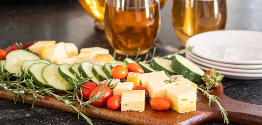 A wooden platter with assorted cheese cubes, cucumber slices, and cherry tomatoes, accompanied by glasses of iced tea and stacked white plates.
