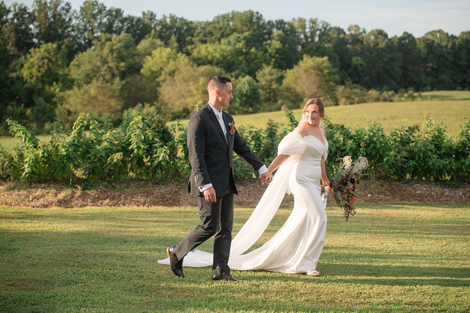 A bride and groom walk hand in hand through a serene, green landscape.