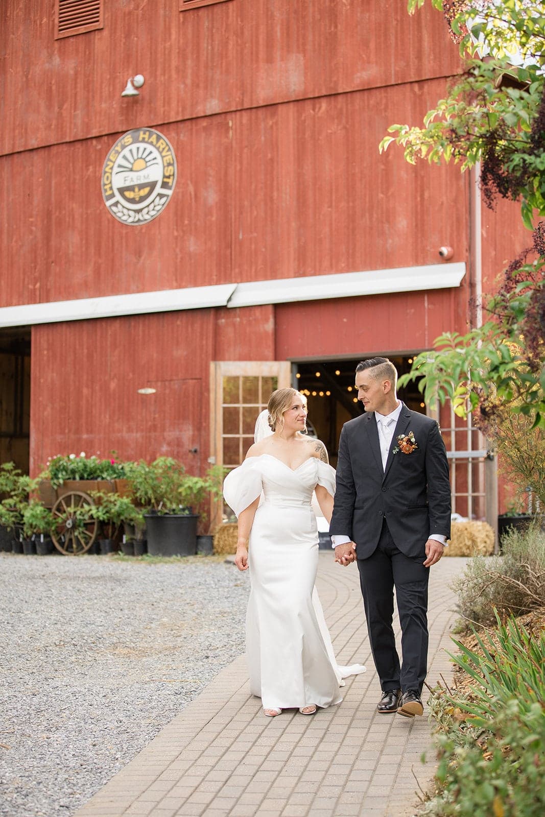 A bride and groom walk hand-in-hand beside a red barn, smiling at each other.