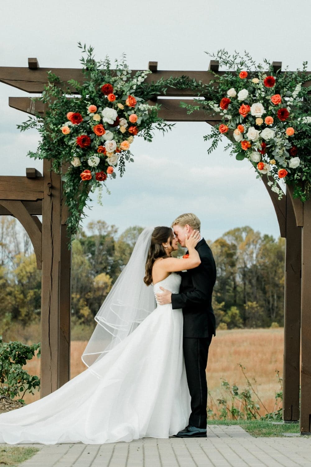 A bride and groom kiss under a floral archway at their outdoor wedding.