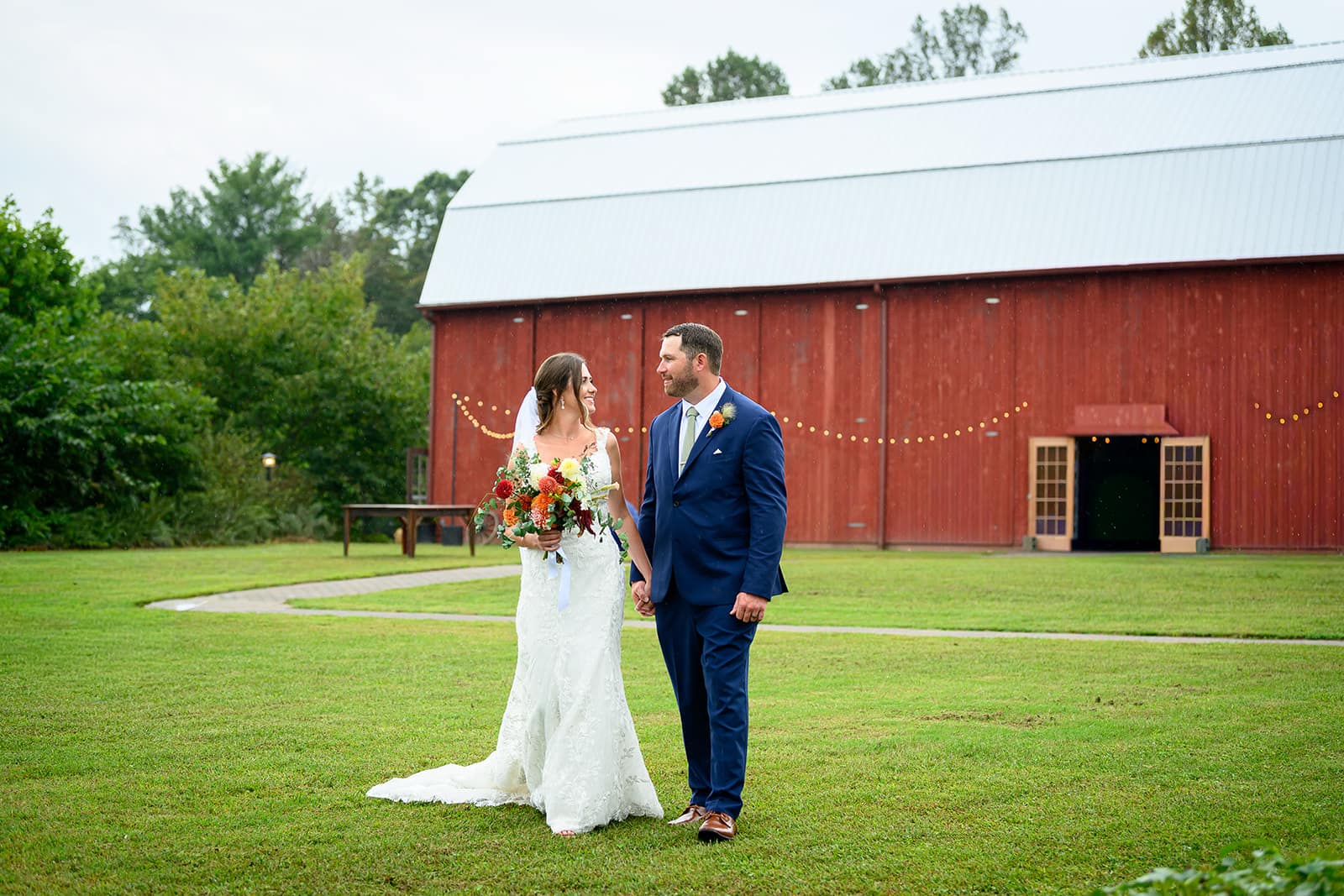 A bride and groom hold hands and smile at each other in front of a rustic red barn.