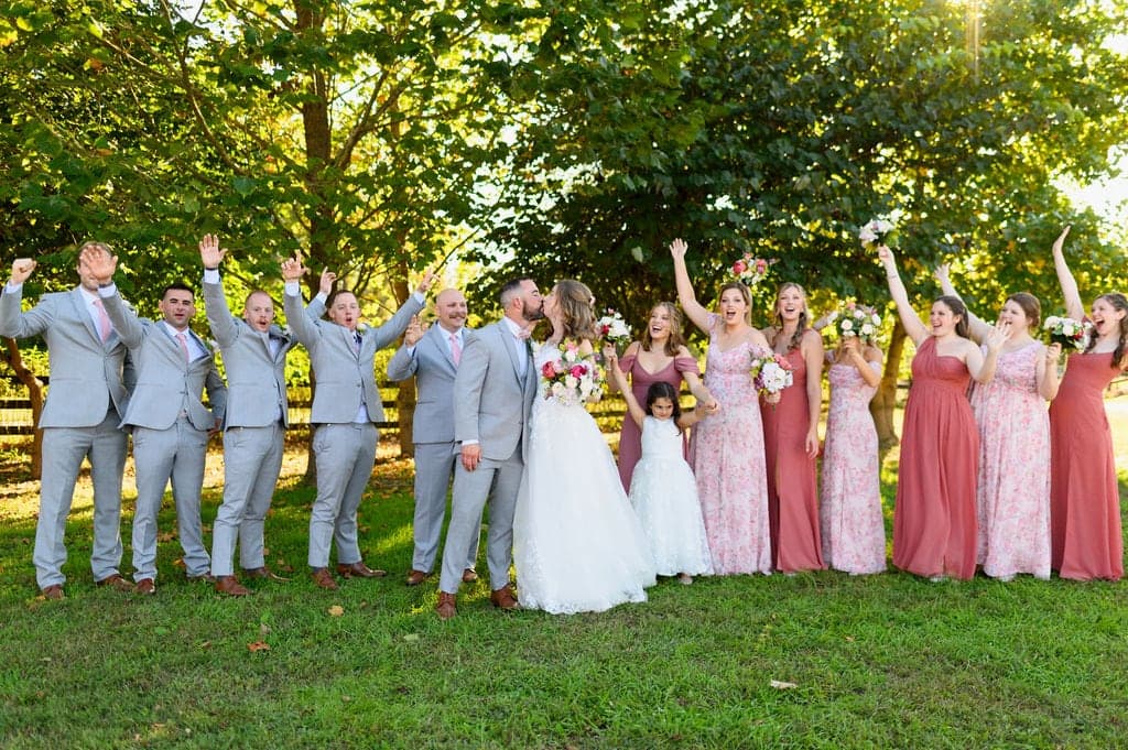 A joyful wedding party celebrates outdoors, with the couple in the center sharing a kiss and guests raising their arms in excitement.