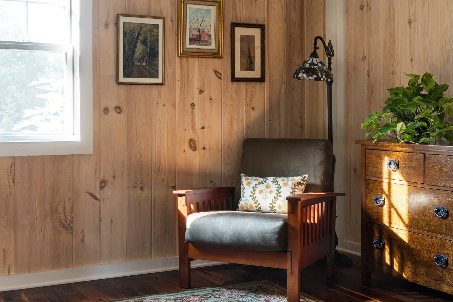 A cozy corner featuring a leather armchair with a decorative pillow beside a lamp, framed artworks on the wall, and a wooden dresser with plants.