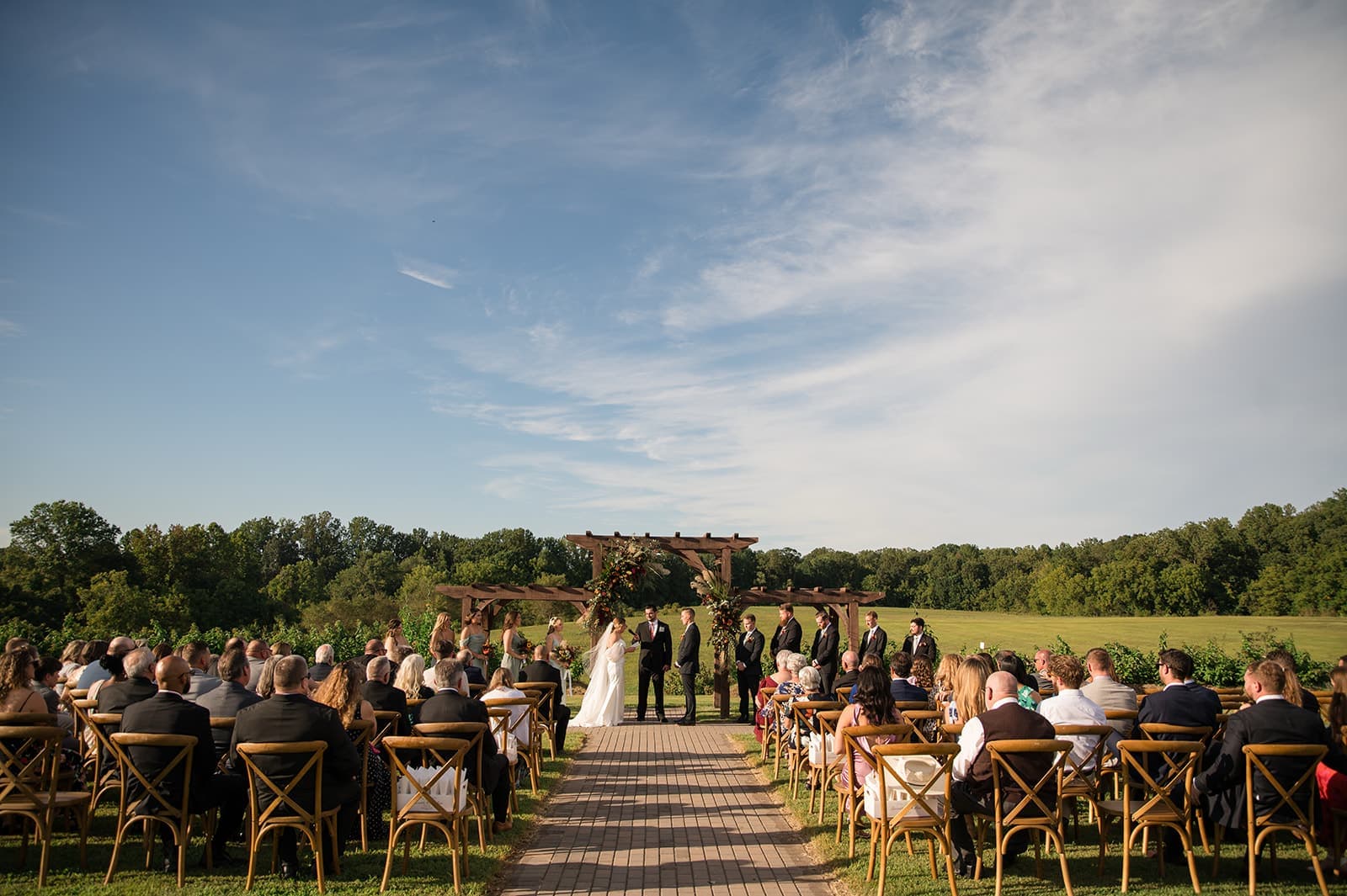 A wedding ceremony taking place outdoors, with guests seated on either side of a wooden pathway.