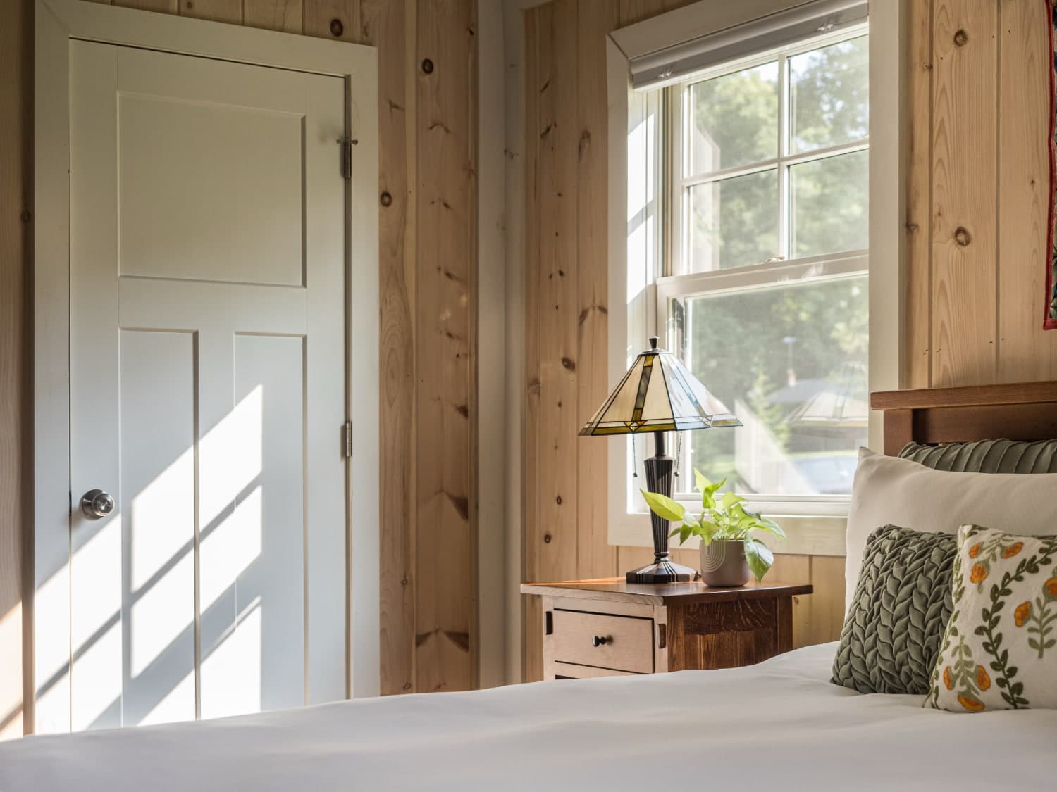 A cozy bedroom featuring a wooden wall, a bed with patterned pillows, a bedside table with a lamp, and a window allowing natural light in.