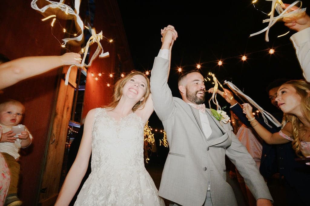 A bride and groom joyfully celebrate their wedding as they raise their hands amidst a festive atmosphere with guests cheering around them.