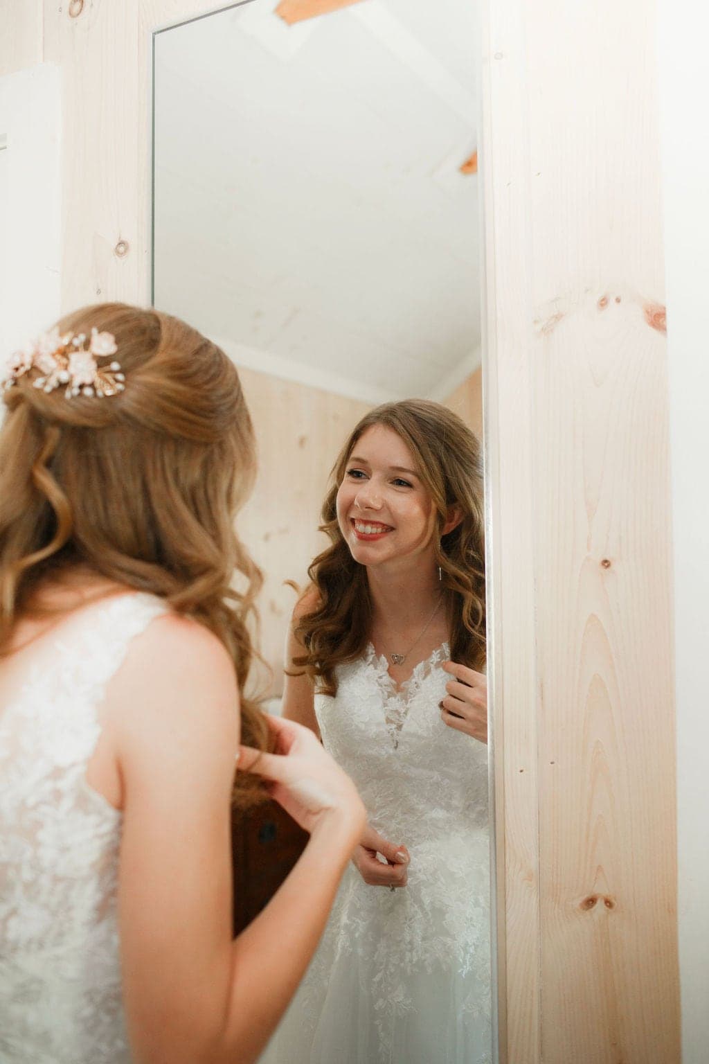 A bride smiles at her reflection in a mirror while adjusting her dress.