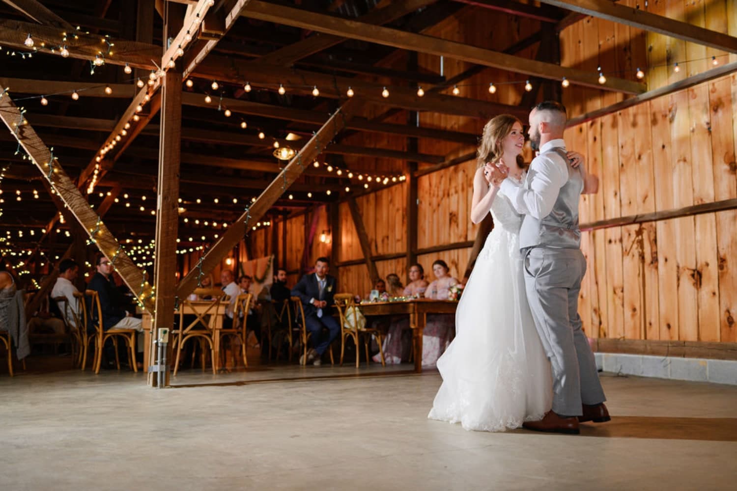 A couple dances together in a rustic barn adorned with string lights, surrounded by seated guests.
