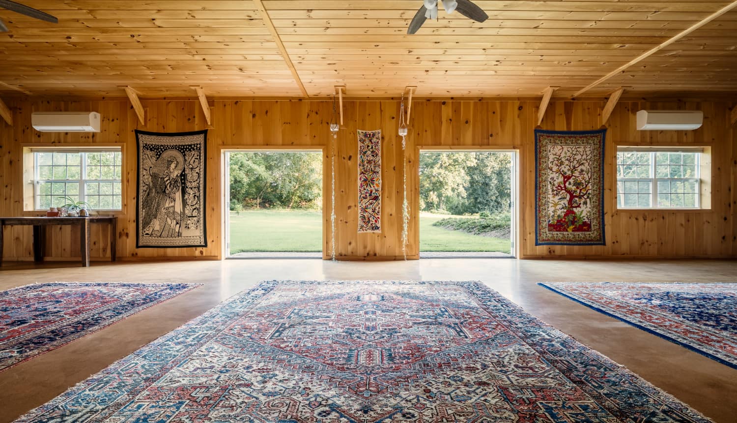 Interior of a wooden room with colorful rugs and tapestry on the walls, opening to a green outdoor view.