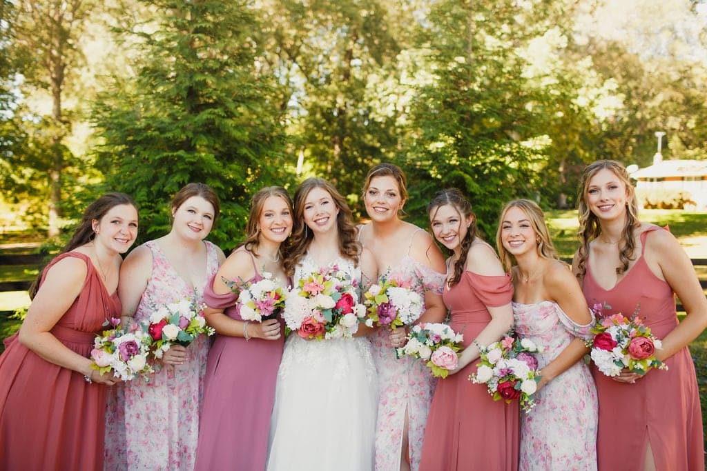 A bride stands with her eight bridesmaids, all smiling and holding bouquets, surrounded by a lush outdoor backdrop.