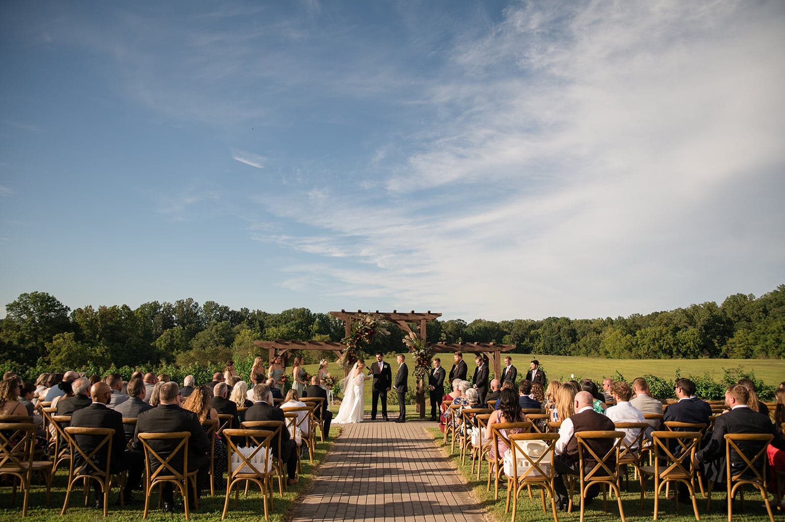 An outdoor wedding ceremony with guests seated on wooden chairs, a couple exchanging vows, and a scenic landscape in the background.