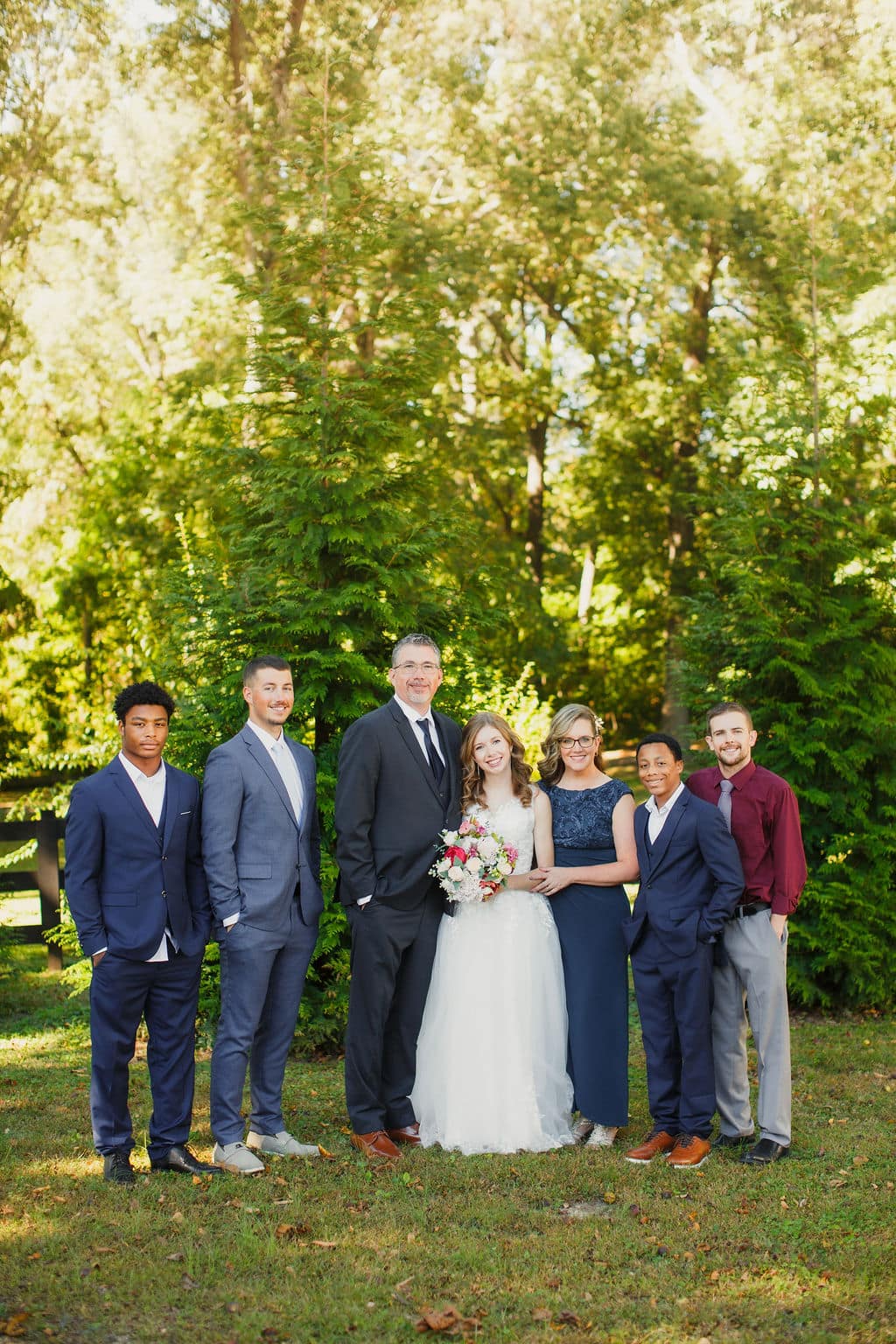 A bride in a white dress stands with six people in formal attire in a green outdoor setting.