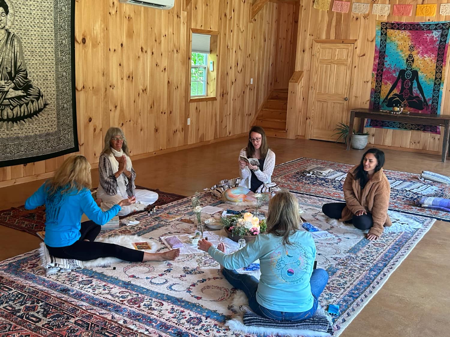 A group of four women engaging in a meditative or spiritual gathering on a patterned rug in a cozy wooden room.