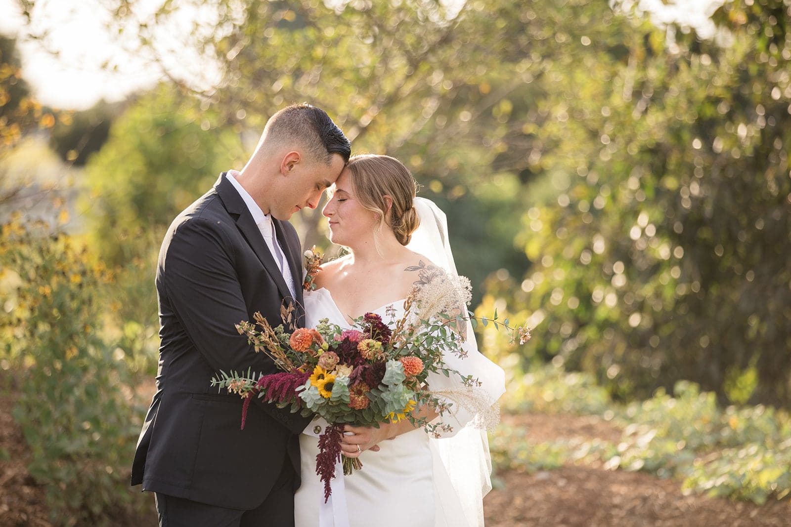A bride and groom share a tender moment outdoors while holding a bouquet.