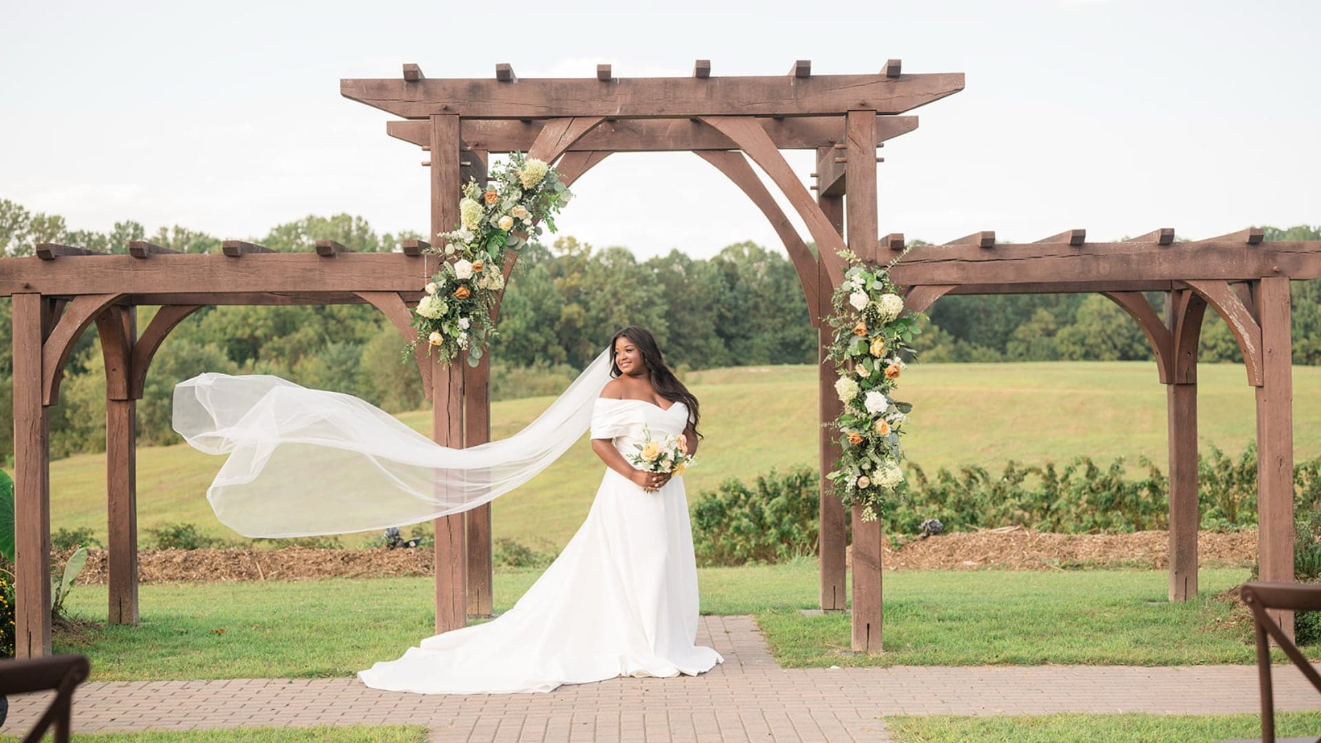 A bride in a flowing white dress stands under a floral arch, with her veil gently blowing in the breeze.