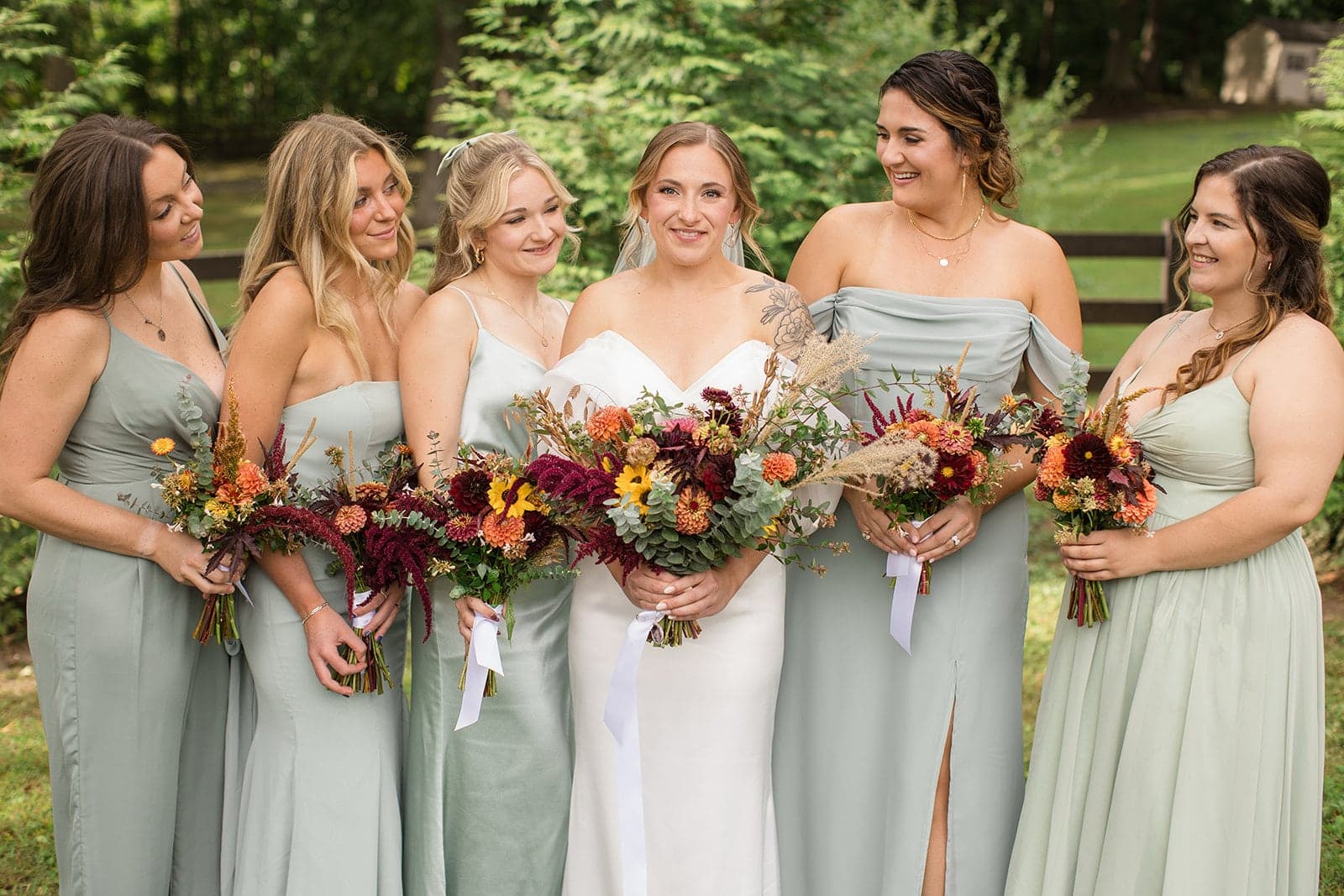 A bride in a white gown poses with her five bridesmaids, all holding floral bouquets, in a lush outdoor setting.