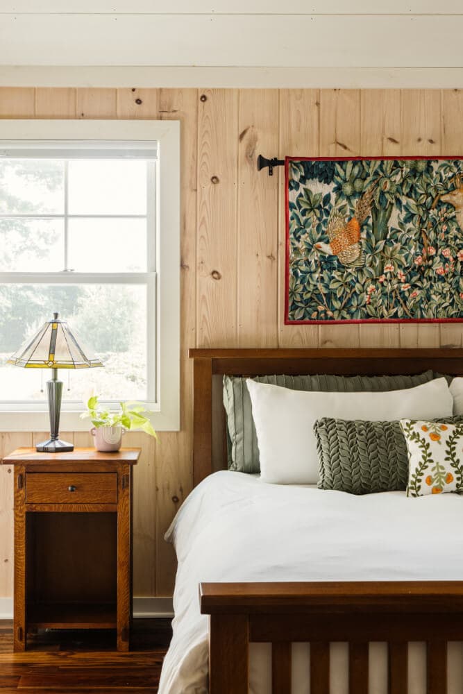 A cozy bedroom featuring a wooden bed, decorative pillows, a bedside lamp, and a colorful wall tapestry.
