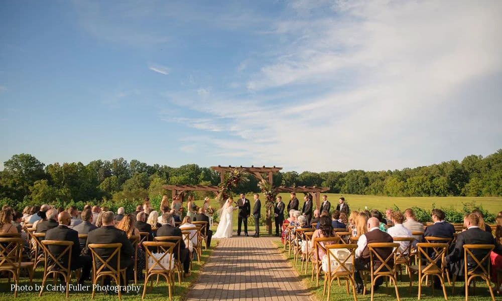 Outdoor wedding ceremony at Honeys Harvest Farm on a lawn with seated guests, a wooden arch, and green farm fields in the background.