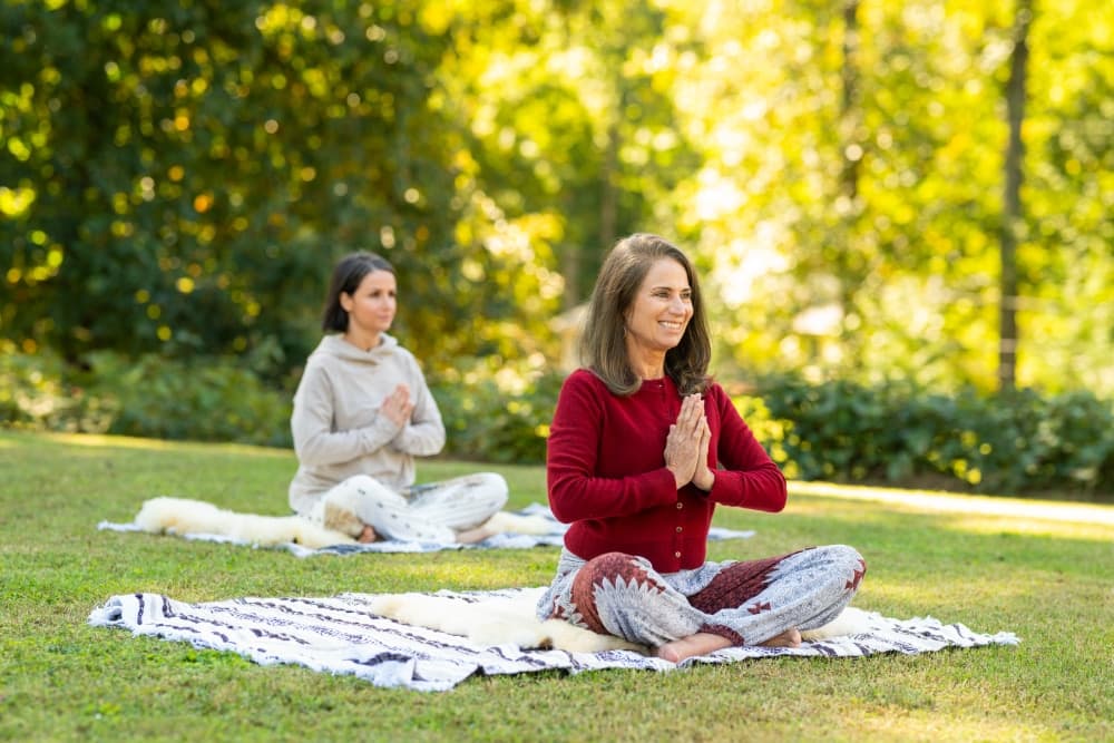 Two women practicing yoga outdoors, seated on mats with hands in a prayer position.