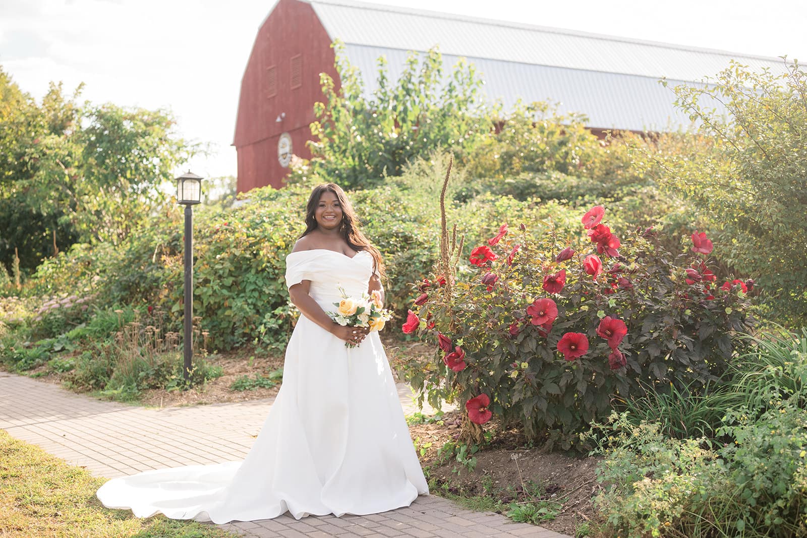 A bride in a white gown stands in a garden with colorful flowers and a red barn in the background.