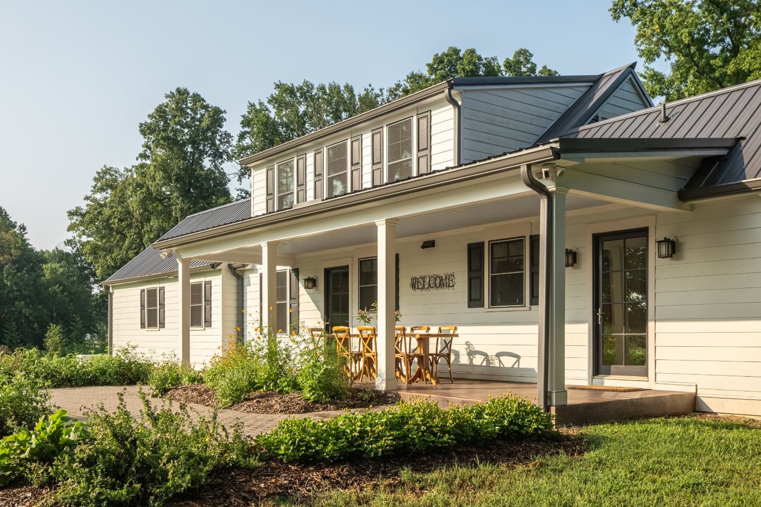 A modern white house with a porch and wooden furniture surrounded by greenery.