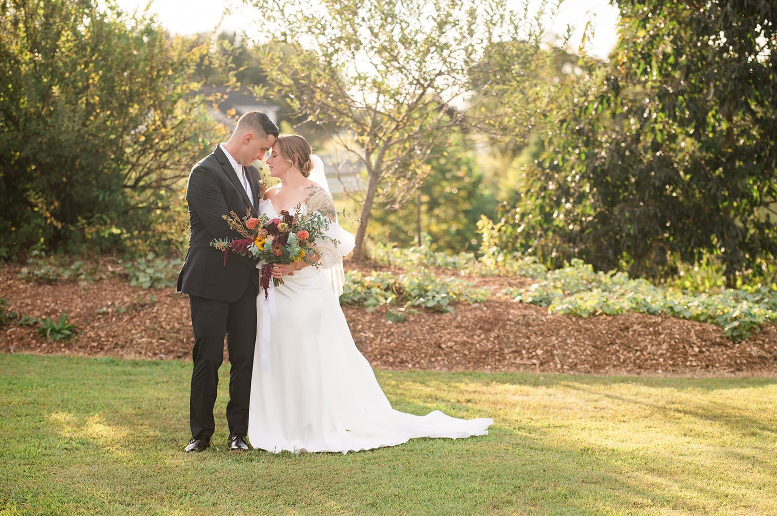A couple shares a tender moment outdoors, surrounded by greenery and holding a bouquet.
