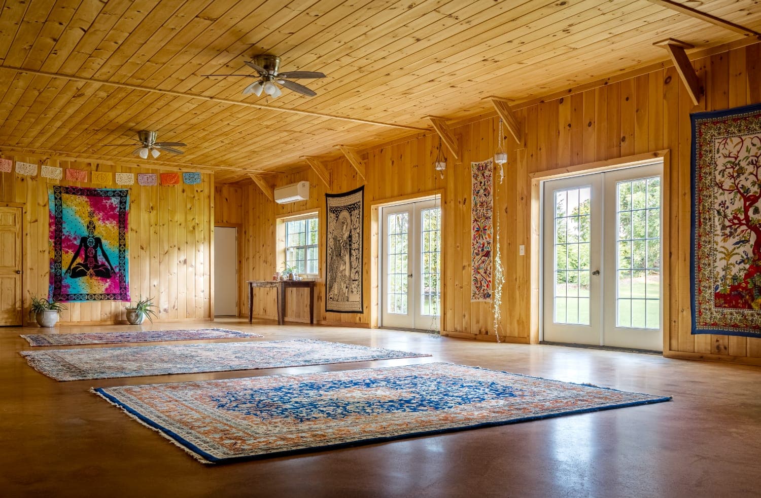 A spacious wooden room with colorful tapestries and patterned rugs, featuring large windows that open to a view.