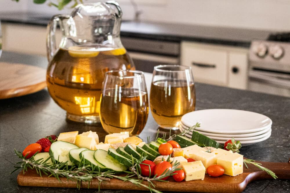 A wooden platter of cheese and vegetables with glasses of iced tea and a pitcher in a kitchen setting.
