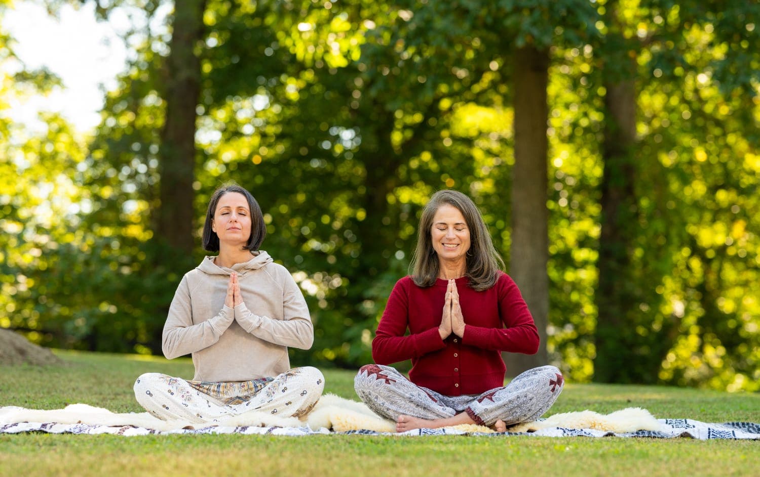 Two women practicing yoga in a tranquil outdoor setting, seated in a meditative pose with hands in prayer.
