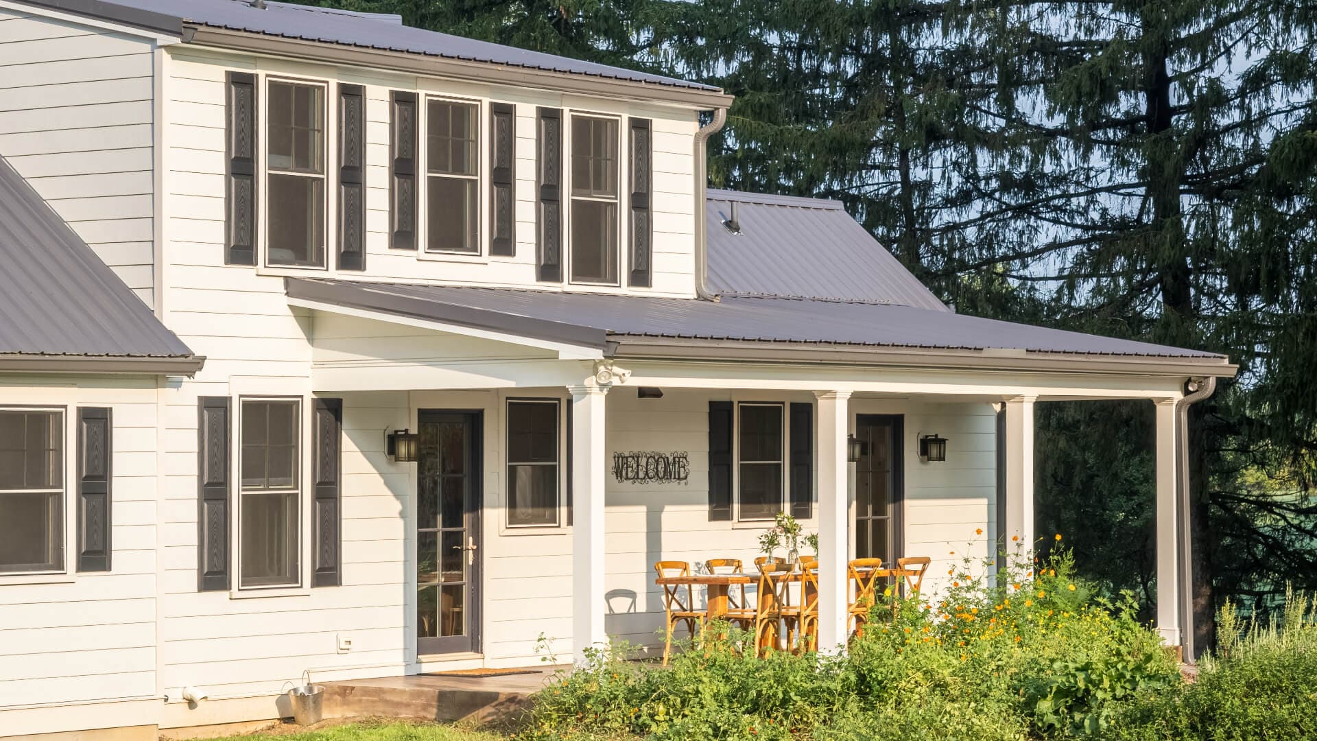 A modern white house with a porch and wooden furniture surrounded by greenery.